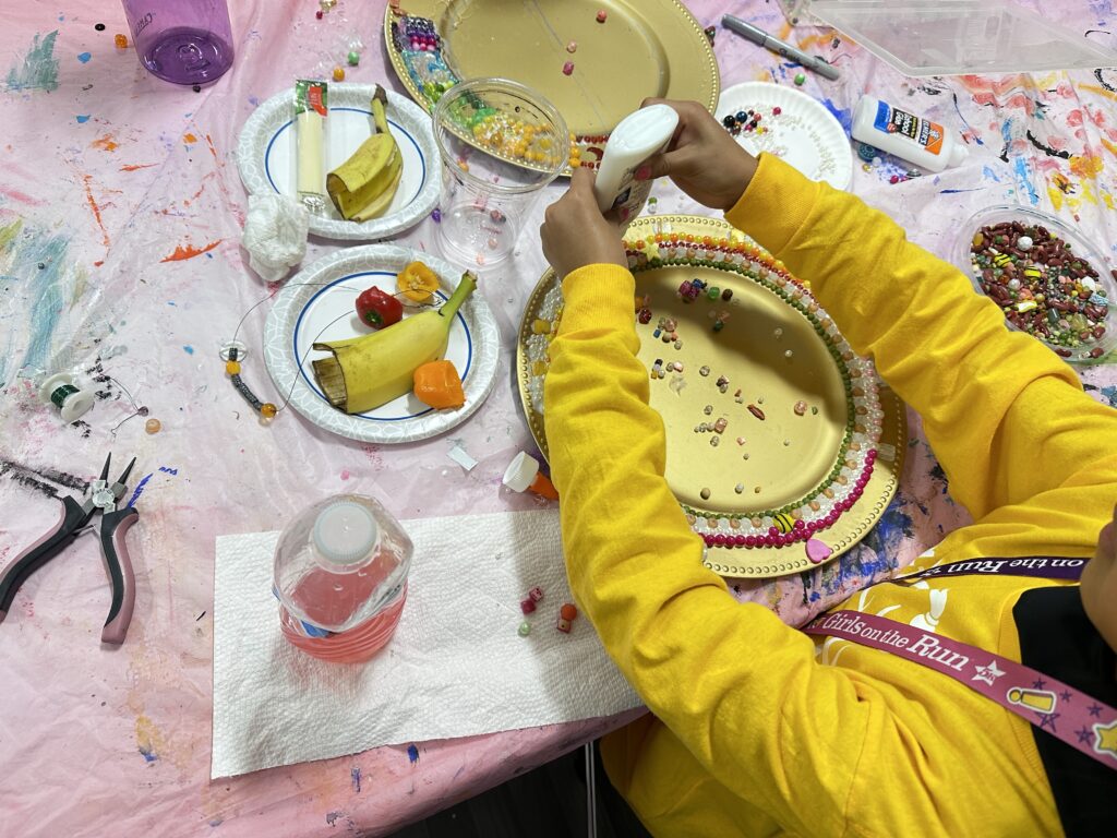 Maria finishes her project, adding glue to the golden plate. She snacks on bell peppers, bananas and a cheese stick provided by the Little Picassos program.