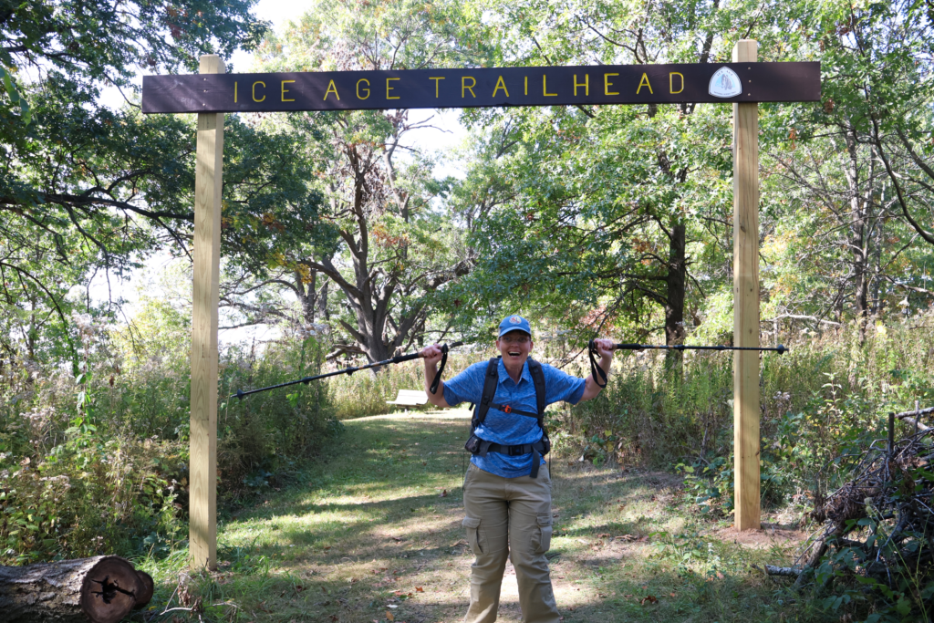 April Scheel smiling with her poles up on the trail while surrounded by trees.
