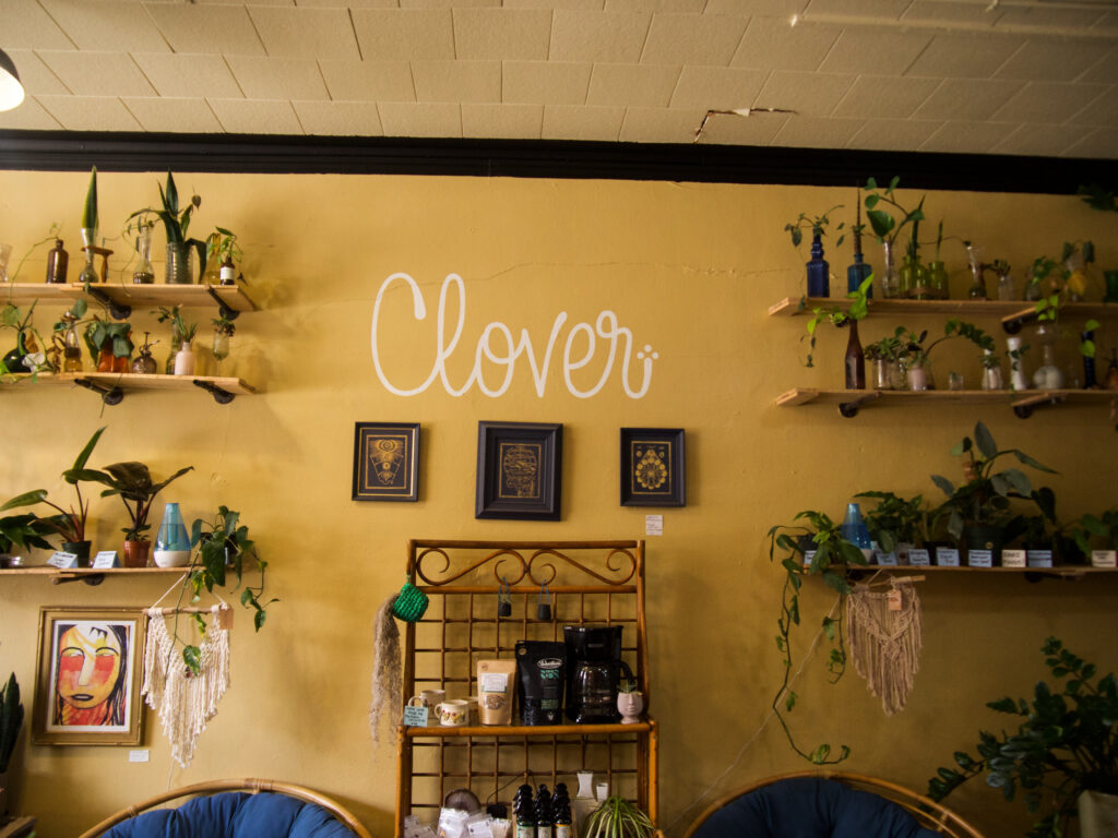 Yellow wall with the text Clover on it, various shelves with plants and other accessories on it. 