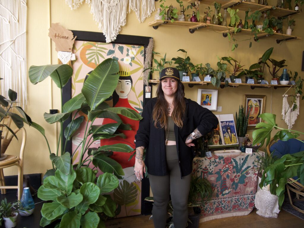 Maggie Murphy standing in her shop surrounded by many large and small plants as well as more colorful pieces of art. 