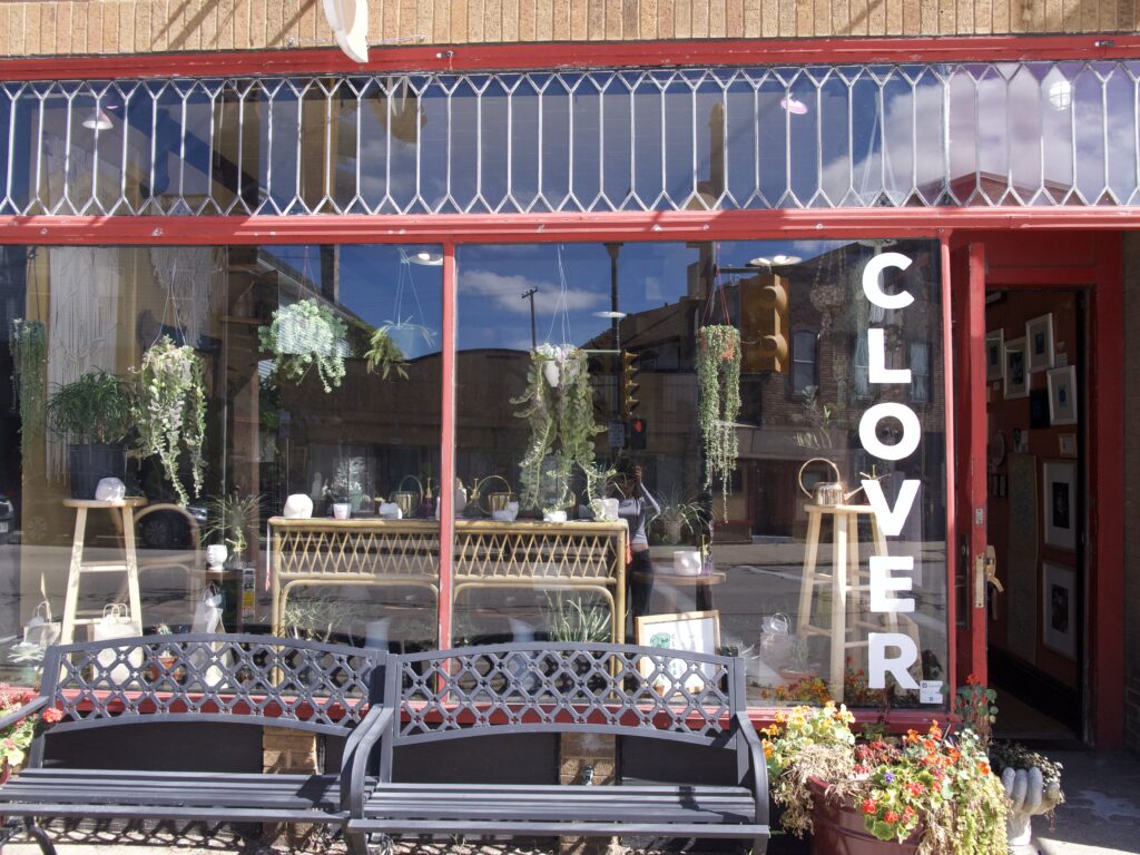 Outside of the Clover plant shop with plants, the stores name, large windows and benches outside. 