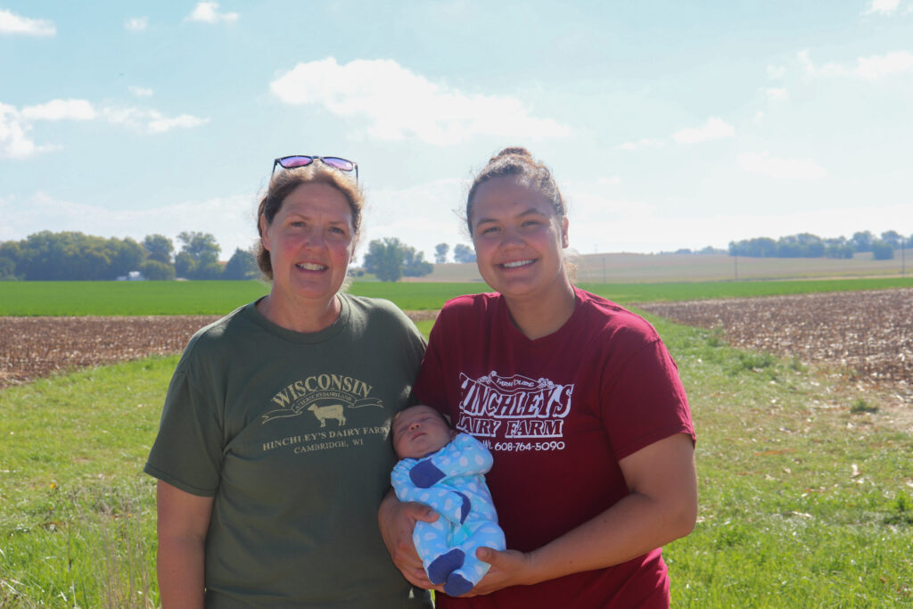 Two women, one holding a baby pose in front of an agricultural field.