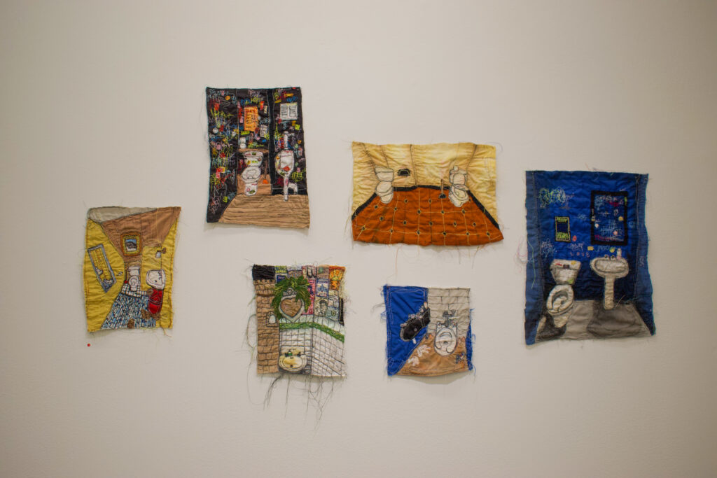 Six embroidered images of toilets and bathrooms displayed on a white wall.