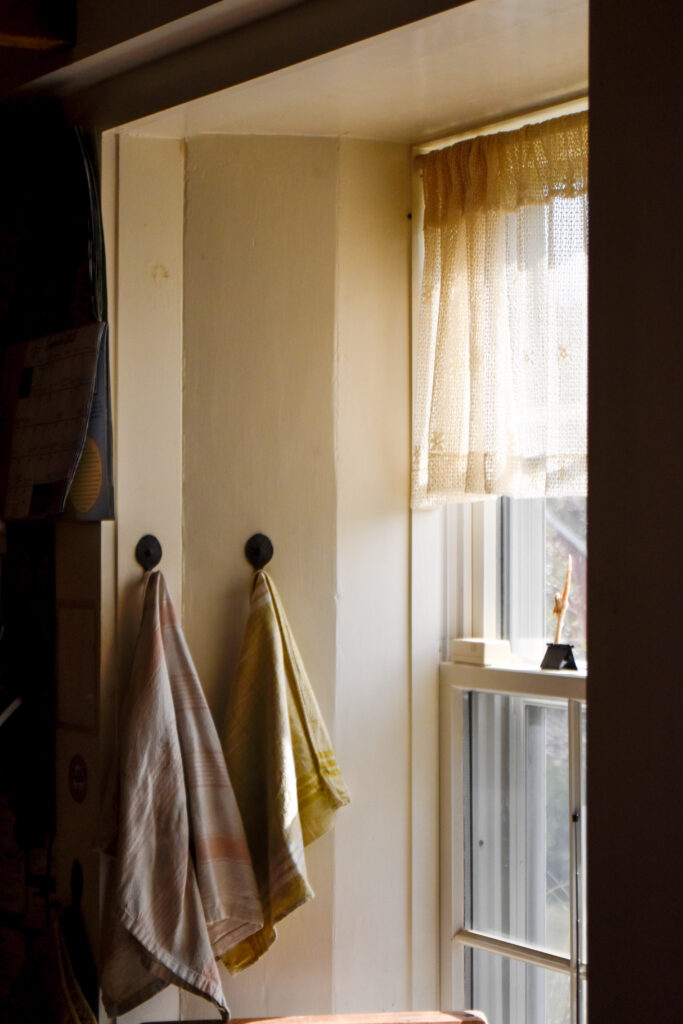 Sunlight streaming into a farmhouse kitchen window with two kitchen towels hanging from hooks. 