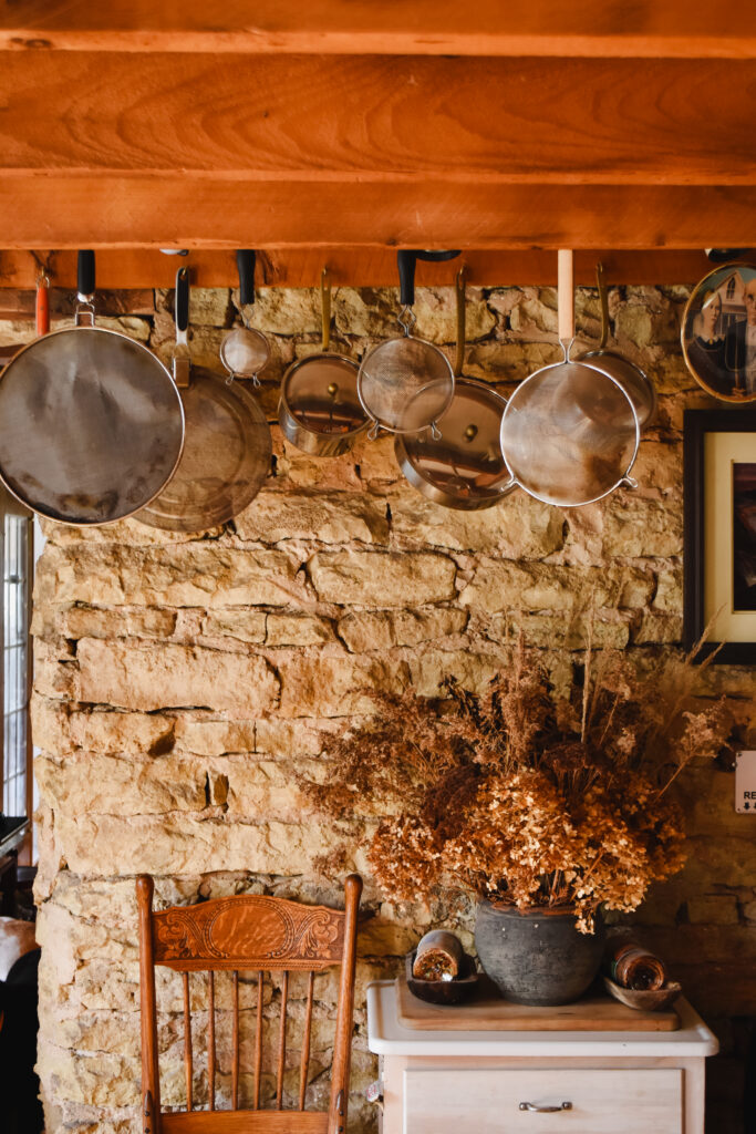 Exposed stone wall of a farmhouse kitchen with pots and pans hanging from the ceiling.