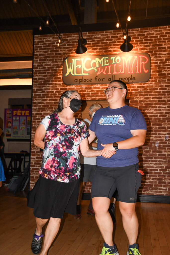 Noelle Gibson stands beside their partner and guides their partner into a turn with a hand behind their back.