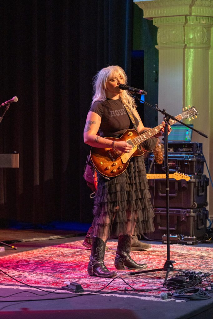 Heyward sings into the microphone, guitar in hand. She wears all black and cowboy boots.