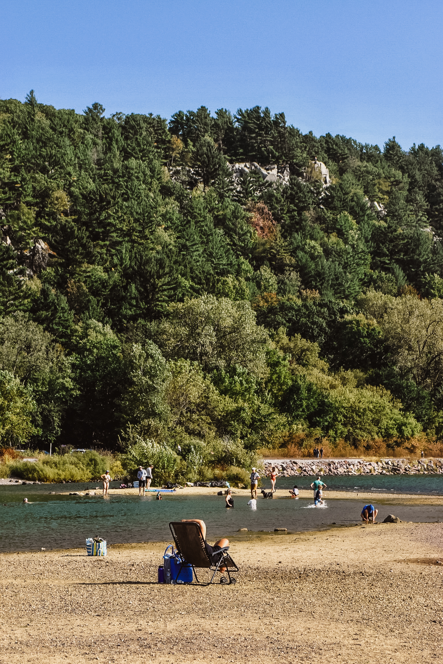 Beach with people swimming and relaxing near a lake and wooded hillside.