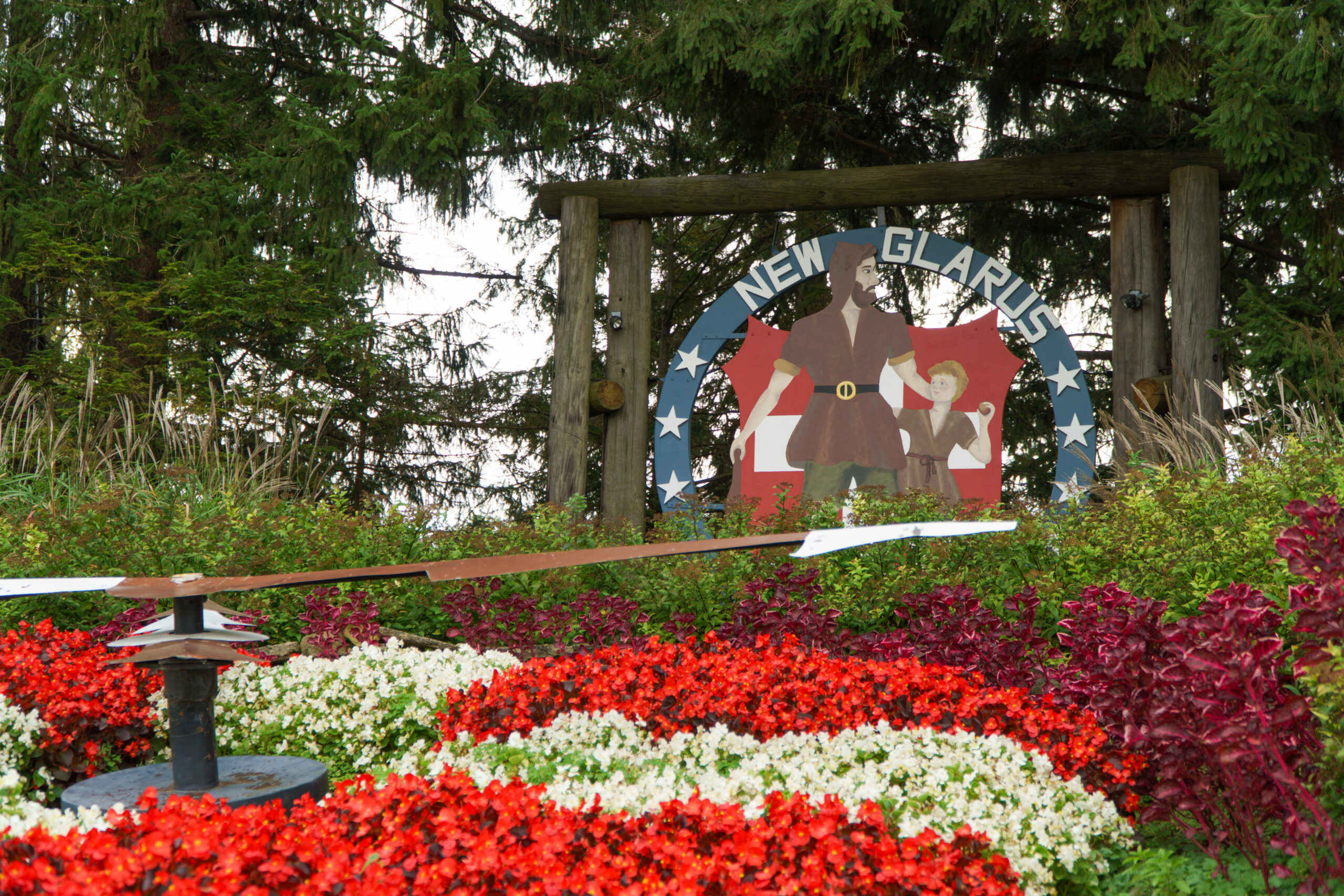 Wooden welcome sign for New Glarus featuring painted figures of a man and child in traditional Swiss-style clothing, surrounded by a circular frame with stars. Bright red, white, and purple flowers fill the foreground, with tall green trees in the background.