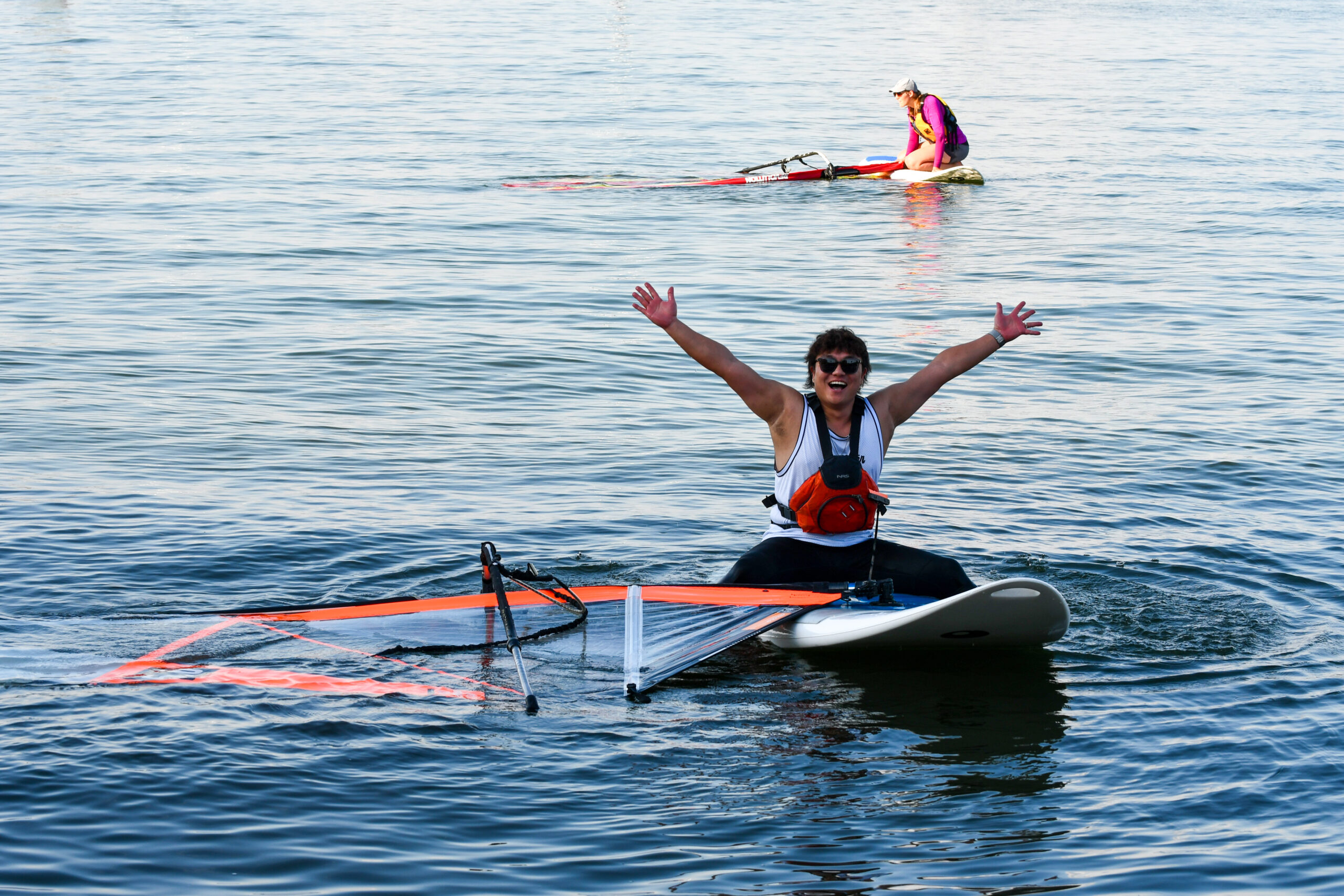 Man holds his arms up to the camera as he sits on his windsurfing board with the sail in the water.